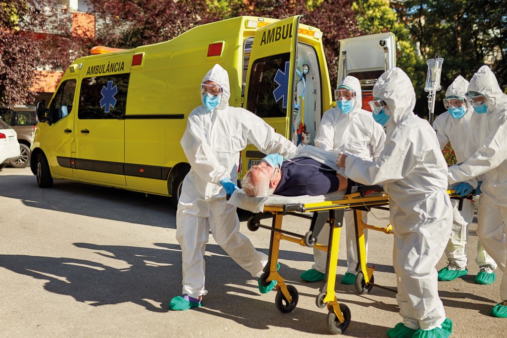 Medical team in protective suits, eyewear, masks, gloves, and shoe protectors wheeling male patient on stretcher with IV drip from ambulance to emergency room.