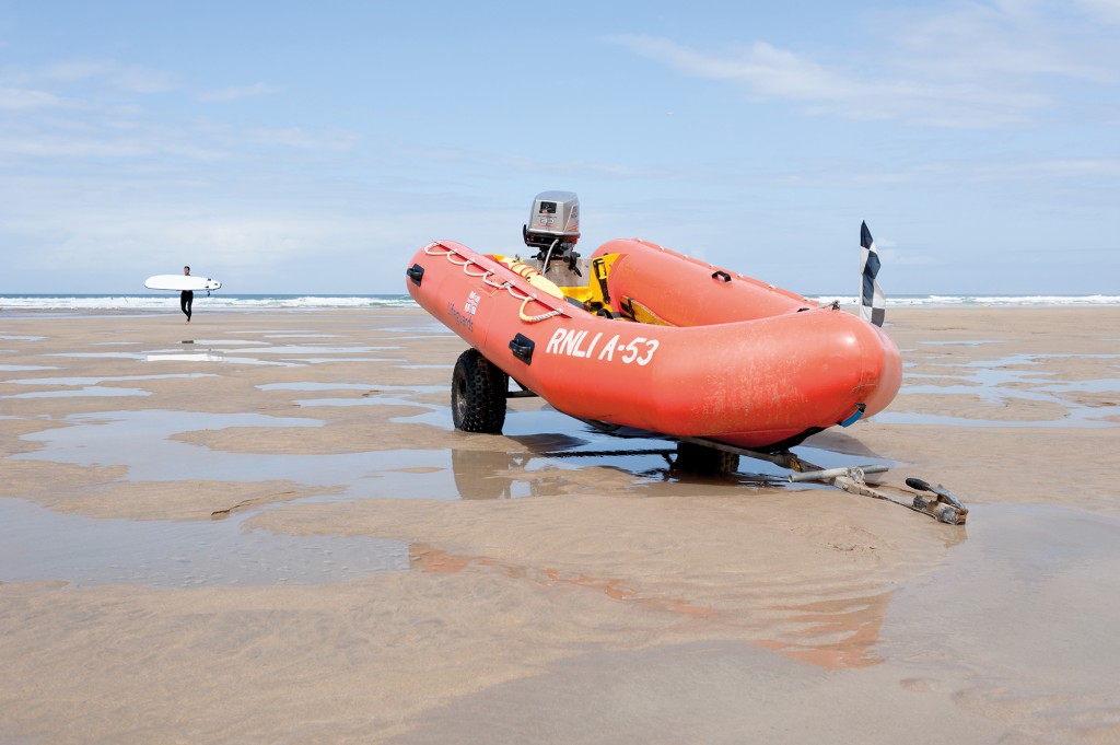 An RNLI inflatable lifeboat resting on a trailer on a Cornish beach with a surfer carrying his surf board in the background.